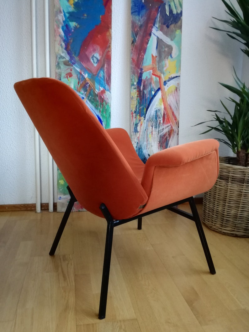 May include: A bright orange velvet armchair with black metal legs. The chair is sitting on a wooden floor.