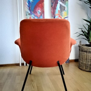 May include: A bright orange armchair with black metal legs. The chair is upholstered in a soft, plush fabric. The chair is sitting on a wooden floor.
