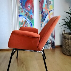 May include: A burnt orange velvet armchair with a black metal frame. The chair has a high back and a wide seat. The chair is sitting on a wooden floor.