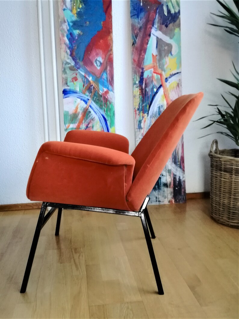 May include: A burnt orange velvet armchair with a black metal frame. The chair has a high back and a wide seat. The chair is sitting on a wooden floor.