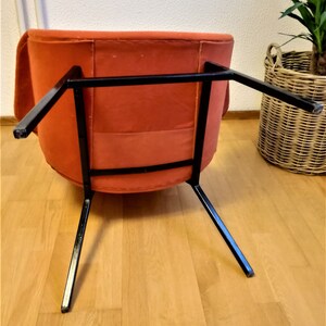 May include: A burnt orange armchair with a black metal frame. The chair is sitting on a wooden floor.