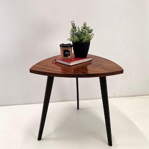May include: A triangular wooden side table with a dark brown finish and three black legs. A small potted plant, a red book, and a candle are on the table's surface. The table is approximately 61 cm wide.
