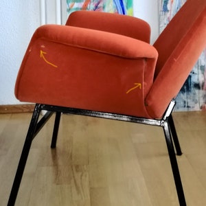 May include: A burnt orange armchair with a black metal frame. The chair has a slightly curved back and a wide seat. The chair is sitting on a wooden floor.