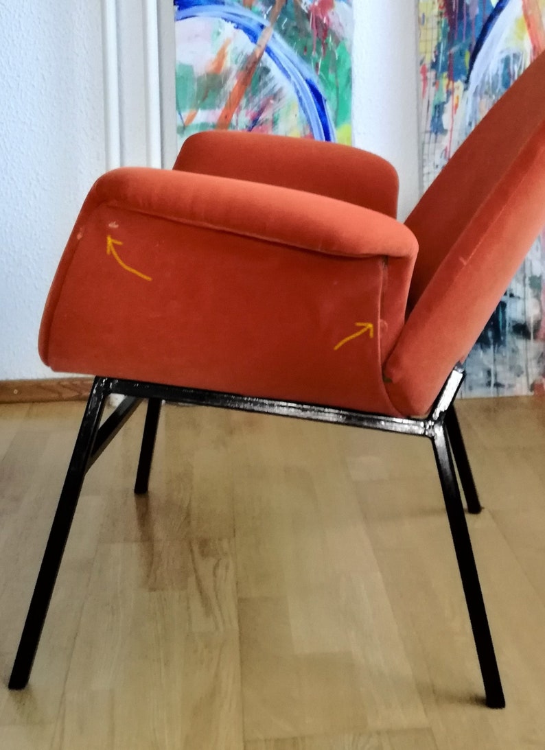 May include: A burnt orange armchair with a black metal frame. The chair has a slightly curved back and a wide seat. The chair is sitting on a wooden floor.