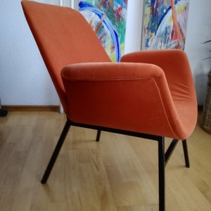 May include: An orange velvet armchair with black metal legs. The chair is angled to show the side profile. The chair is in front of a white wall with two abstract paintings.