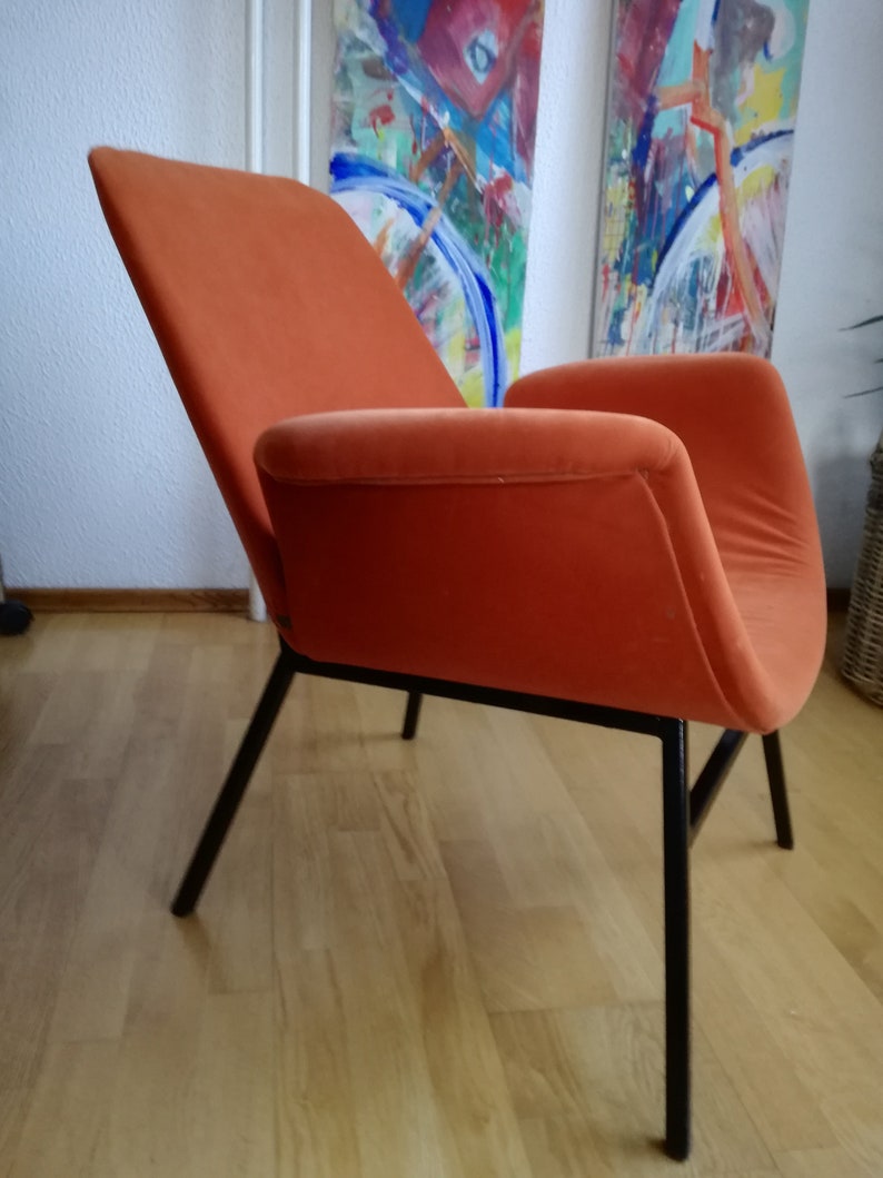May include: An orange velvet armchair with black metal legs. The chair is angled to show the side profile. The chair is in front of a white wall with two abstract paintings.