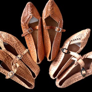 May include: Several pairs of handmade leather shoes in a tan hue, with a pointed toe and embossed detailing. Each shoe has a buckle strap for adjustment. The shoes are displayed against a black backdrop.