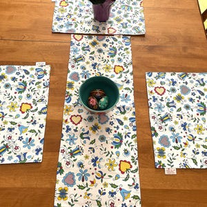 May include: Three white table runners with a floral pattern in shades of blue, yellow, and red. The runners are placed on a wooden table with a small green bowl filled with colorful Easter eggs in the center.