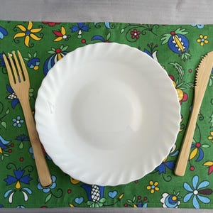 Puede incluir: Un plato de cerámica blanco con un borde ondulado se encuentra sobre un mantel individual verde con un patrón floral colorido. Un tenedor y un cuchillo de madera están colocados sobre el mantel individual.