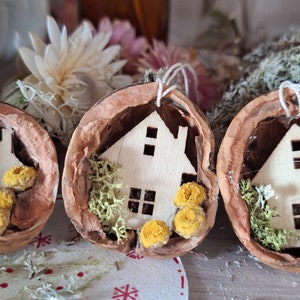 Könnte beinhalten: Drei Walnussschalen-Ornamente mit einem kleinen, ausgeschnittenen Holzhäuschen in jeder. Die Häuser sind weiß gestrichen und haben kleine Fenster. Jedes Ornament ist mit grünem Moos und gelben Blumen verziert.