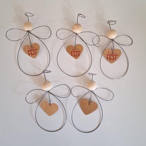 Könnte beinhalten: Fünf handgefertigte Engel-Ornamente. Jeder Engel hat einen Drahtrahmen, einen Holzkopf und ein herzförmiges Etikett mit dem roten Text "frohes FEST". Die Flügel werden durch geschwungenen Draht gebildet.