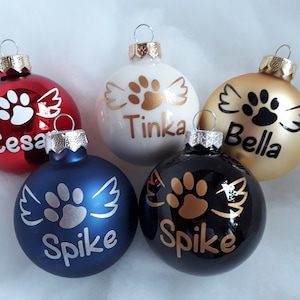 Könnte beinhalten: Fünf Weihnachtskugeln mit einem Pfotenabdruck- und Flügeldesign. Die Kugeln sind rot, weiß, gold, blau und schwarz. Jede Kugel hat einen anderen Namen: Cesa, Tinka, Bella, Spike und Spike.