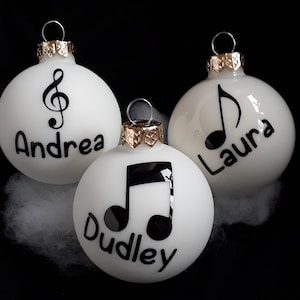 Könnte beinhalten: Drei weiße Weihnachtskugeln mit schwarzen Noten und den Namen "Andrea", "Laura" und "Dudley" darauf.