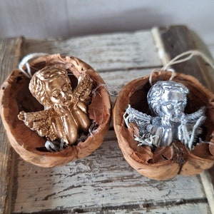 Könnte beinhalten: Zwei kleine Engelsfiguren, eine goldene und eine silberne, befinden sich in zwei Walnussschalen. Die Schalen werden als Dekoration verwendet.