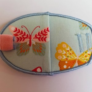 May include: A light blue eye patch with a butterfly design. The patch has a pink fabric strap and a blue embroidered border.