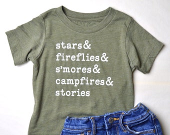 Op de afbeelding: Een groen T-shirt met korte mouwen met de tekst "stars & fireflies & s'mores & campfires & stories" in wit gedrukt. Het shirt wordt gecombineerd met een blauwe denim short.