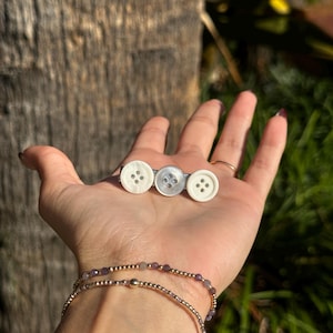 May include: A hair clip featuring three white buttons with four holes each, attached to a silver bar. The buttons are in varying shades of white. The clip is held in a hand, with two gold bracelets with purple beads on the wrist.