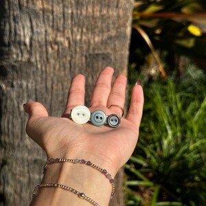 May include: Three buttons in a row, in white, teal, and dark gray. The buttons have two holes each. A gold bracelet with purple beads and a gold bracelet with a single gold bead are also visible.