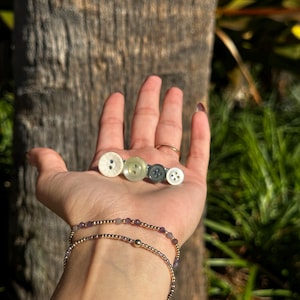 May include: A collection of four decorative hair clips, each featuring a unique button design in shades of white, green, and gray. The clips are displayed on a hand, with a gold beaded bracelet visible on the wrist. The background is a blurred outdoor scene.