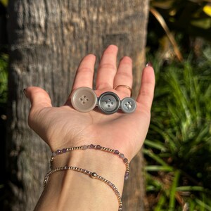 May include: Three buttons in varying shades of gray are held in a hand, with a gold bracelet and a beaded bracelet with purple beads. The buttons are round with four holes each. The background is a blurred view of a tree trunk and green foliage.