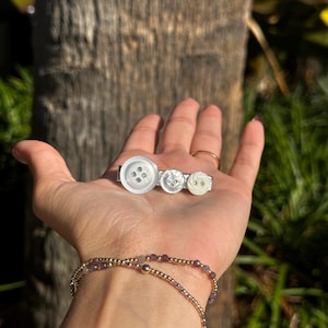 May include: A handmade barrette featuring three white buttons of varying sizes. The buttons are attached to a silver-toned metal barrette. A gold and amethyst beaded bracelet is also visible.