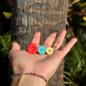 May include: Three colorful buttons: red, turquoise, and yellow. The red button is the largest, followed by the turquoise and then the yellow. Each button has four holes. The buttons are displayed on a hand.