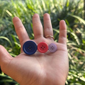 May include: Three colorful buttons: a large blue button, a medium red button, and a small purple button. Each button has four holes. The buttons are arranged in a row, suggesting a decorative or craft item.