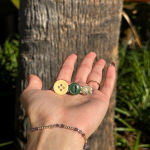 May include: Three button rings in a hand. The rings feature a light yellow button, a dark green button, and a clear button with a white center. The rings are set on silver bands. A gold beaded bracelet with purple beads is also visible.