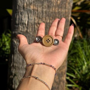 May include: A collection of buttons and bracelets. The buttons include a large, light brown button with four holes and two smaller, silver-toned buttons with decorative designs. Two delicate gold bracelets with small beads are also visible.