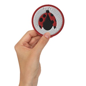 May include: A red and black embroidered ladybug patch with a white background. The patch is circular and has a red border.