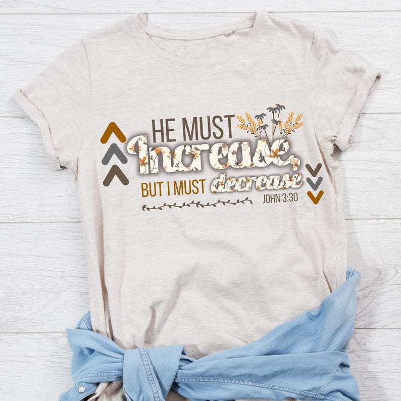 May include: A light beige t-shirt featuring the text "HE MUST increase BUT I MUST decrease JOHN 3:30". The text is in various fonts and colours, with floral details. A denim jacket is tied around the waist.