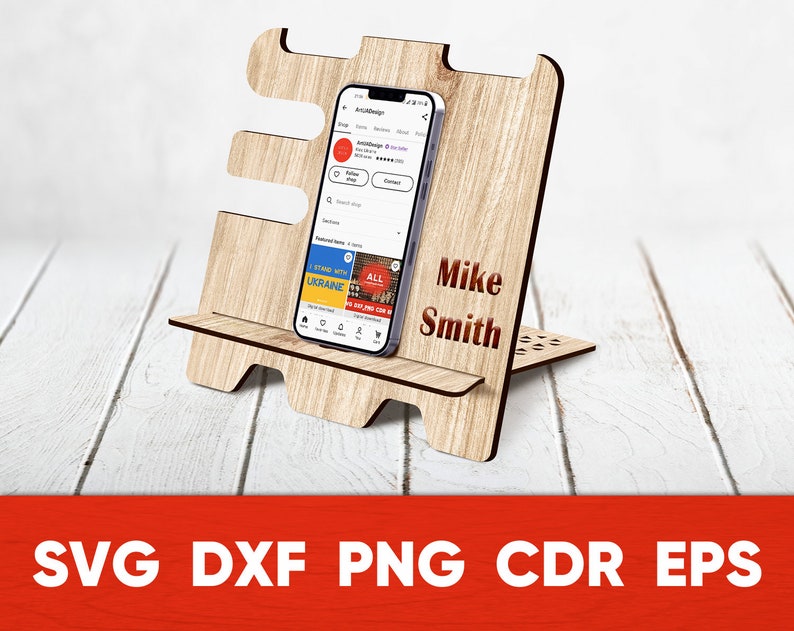 May include: A wooden phone stand with a personalized name, "Mike Smith", engraved on the front. The stand is designed to hold a smartphone upright, with a small shelf for the phone to rest on. The stand is made of light brown wood and has a natural wood finish.