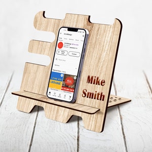 May include: A wooden phone stand with a personalized name, "Mike Smith", engraved on the front. The stand is designed to hold a smartphone upright, and has a cutout for charging cables. The stand is made of light brown wood and has a natural wood finish.