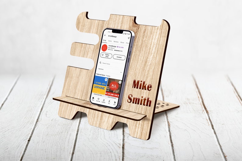May include: A wooden phone stand with a personalized name, "Mike Smith", engraved on the front. The stand is designed to hold a smartphone upright, and has a cutout for charging cables. The stand is made of light brown wood and has a natural wood finish.