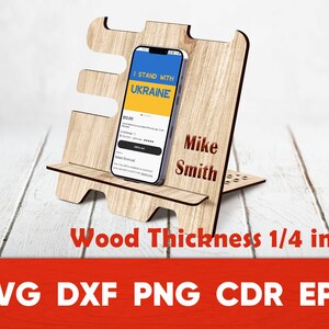 May include: A wooden phone stand with a cutout design. The stand is 1/4 inch thick and has a slot for a phone. The phone in the stand displays the text "I Stand With Ukraine". The stand is personalized with the name "Mike Smith".