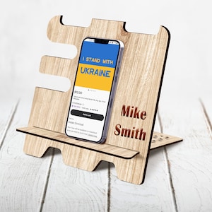 May include: A wooden phone stand with a cutout design. The stand has a small shelf for a phone and a larger shelf for a tablet. The stand is personalized with the name "Mike Smith". The phone screen shows a blue and yellow app with the text "I Stand With Ukraine".