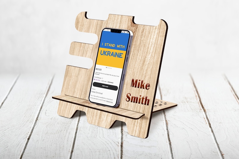 May include: A wooden phone stand with a cutout design. The stand has a small shelf for a phone and a larger shelf for a tablet. The stand is personalized with the name "Mike Smith". The phone screen shows a blue and yellow app with the text "I Stand With Ukraine".