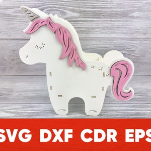 May include: A white wooden unicorn-shaped organizer with pink accents. The unicorn has a horn, mane, and tail, all in pink. The design includes cut-out stars and slots. The text "SVG DXF CDR EPS" is at the bottom.