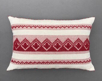 May include: A white rectangular pillow with a red embroidered pattern. The pattern features a repeating design of stylized hearts and geometric shapes.