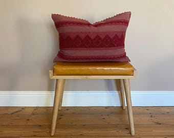 May include: A decorative red pillow with a geometric pattern rests on a small wooden bench with a yellow leather seat. The pillow has a textured design and is placed in front of a light-colored wall and wooden floor.