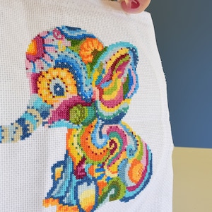 May include: A colorful cross-stitch embroidery of an elephant with a paisley pattern. The elephant is stitched in shades of blue, green, yellow, orange, pink, and purple.