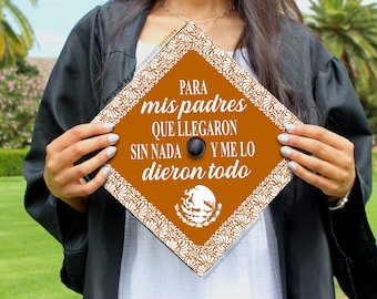 Puede incluir: Un gorro de graduación con fondo marrón y borde floral blanco. El texto en el gorro dice "Para mis padres que llegaron sin nada y me lo dieron todo". El gorro también presenta una imagen estilizada del escudo de armas mexicano.