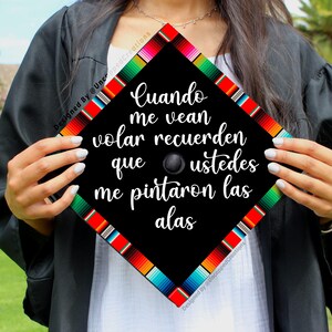 May include: A black graduation cap with a colorful Mexican serape design. The cap has white text that reads "Cuando me vean volar recuerden que ustedes me pintaron las alas".