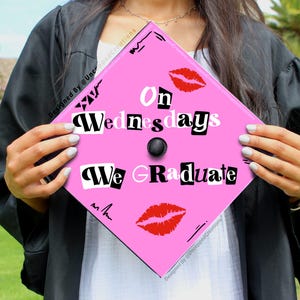 May include: Pink graduation cap with the text "On Wednesdays We Graduate" in black and white block letters. The text is styled to look like magazine cutouts. There are two red lips printed on the cap.
