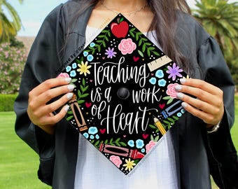 May include: A black graduation cap decorated with colorful floral and school-themed illustrations. The cap features the text "Teaching is a work of Heart" in white script, surrounded by an apple, crayons, and flowers.