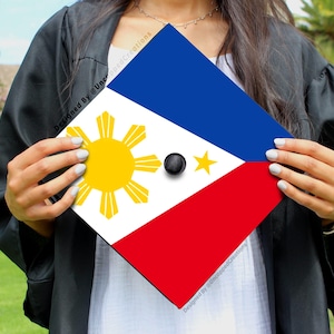 May include: A graduation cap decorated with the flag of the Philippines. The cap is blue, white, and red with a yellow sun and a star in the center. The cap is being held by a person wearing a black graduation gown.