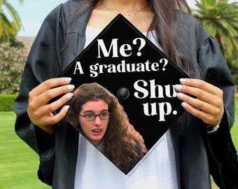 May include: Black graduation cap with the text "Me? A graduate? Shut up." and a photo of a woman wearing glasses. The cap is held by a person wearing a graduation gown, with a green lawn and palm trees in the background.
