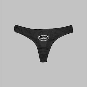 May include: Black thong underwear featuring a white oval graphic on the front center. Inside the oval, the word hysteric is printed in a white cursive script, with small white spikes extending from the top edge of the oval border.
