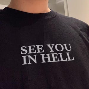 May include: A black t-shirt featuring the white text "SEE YOU IN HELL" printed in a bold, serif font across the chest area. The garment has a standard crew neckline and is constructed from a solid black fabric with a smooth, matte finish.