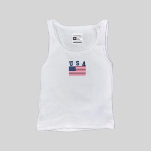 May include: White ribbed vest top featuring the word "USA" in navy blue block letters and a small American flag graphic. The vest top has a scoop neck and a tag at the top.